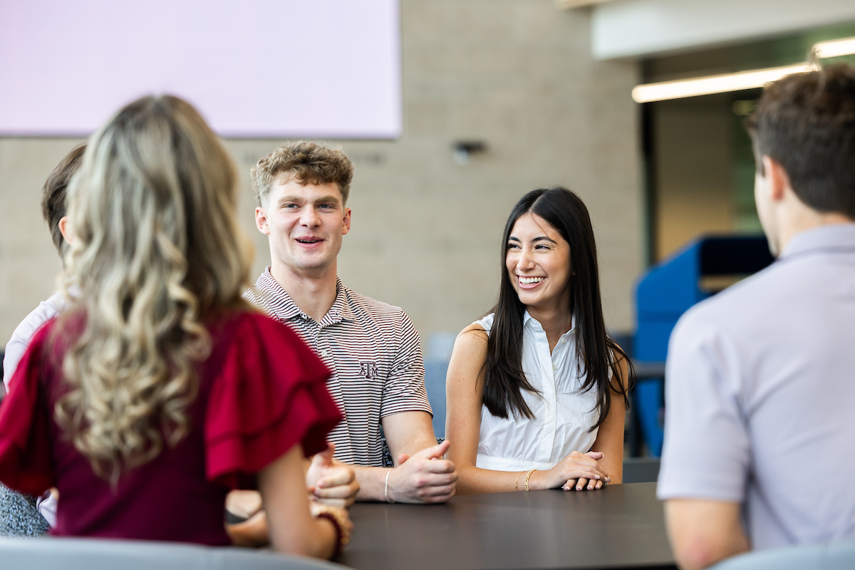 Five Texas A&M students sit facing each other at a table, smiling and talking.