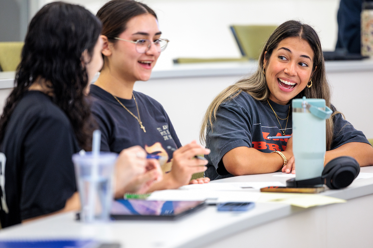 Three Texas A&M students sit in a classroom smiling and talking with course materials on the desk in front of them.